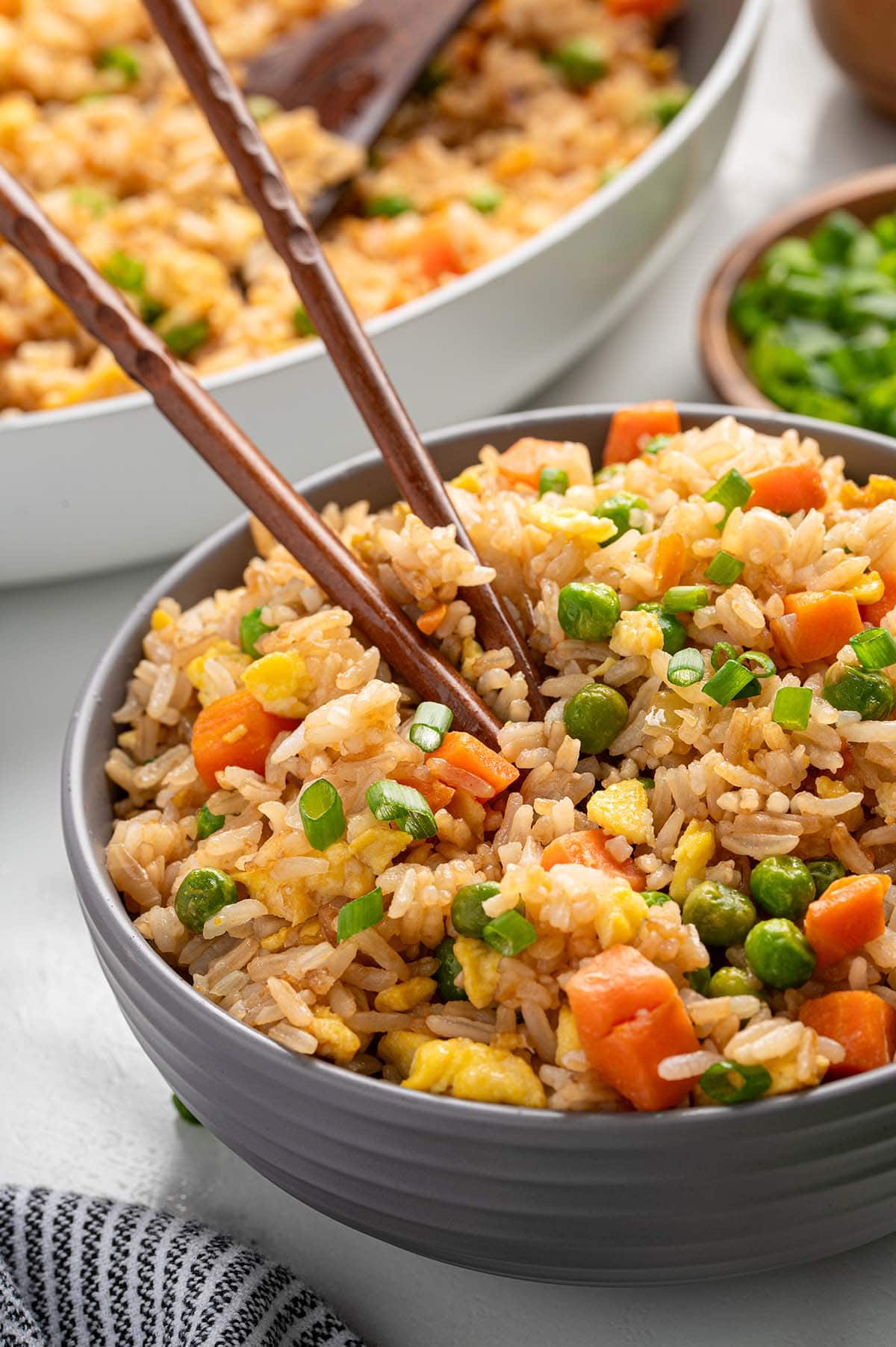 Bowl of homemade fried rice with carrots, peas, scrambled eggs, and green onions, served with chopsticks and a skillet of fried rice in the background.