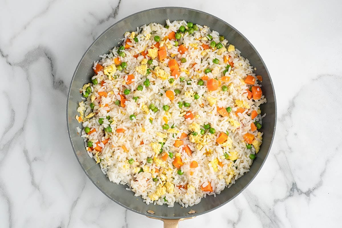 Overhead view of white rice mixed with peas, carrots, scrambled eggs, and onions in a skillet before adding sauce for homemade fried rice.
