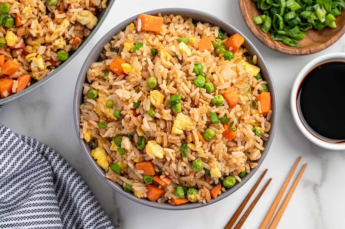 Bowl of easy homemade fried rice with carrots, peas, scrambled eggs, onions, and green onions, served with soy sauce and chopsticks on the side.
