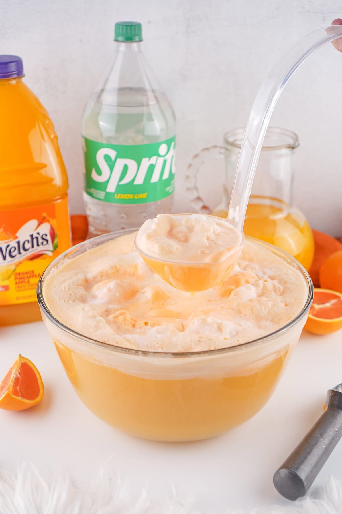 Ladle serving creamy orange punch from a glass bowl with citrus drinks and orange slices nearby.