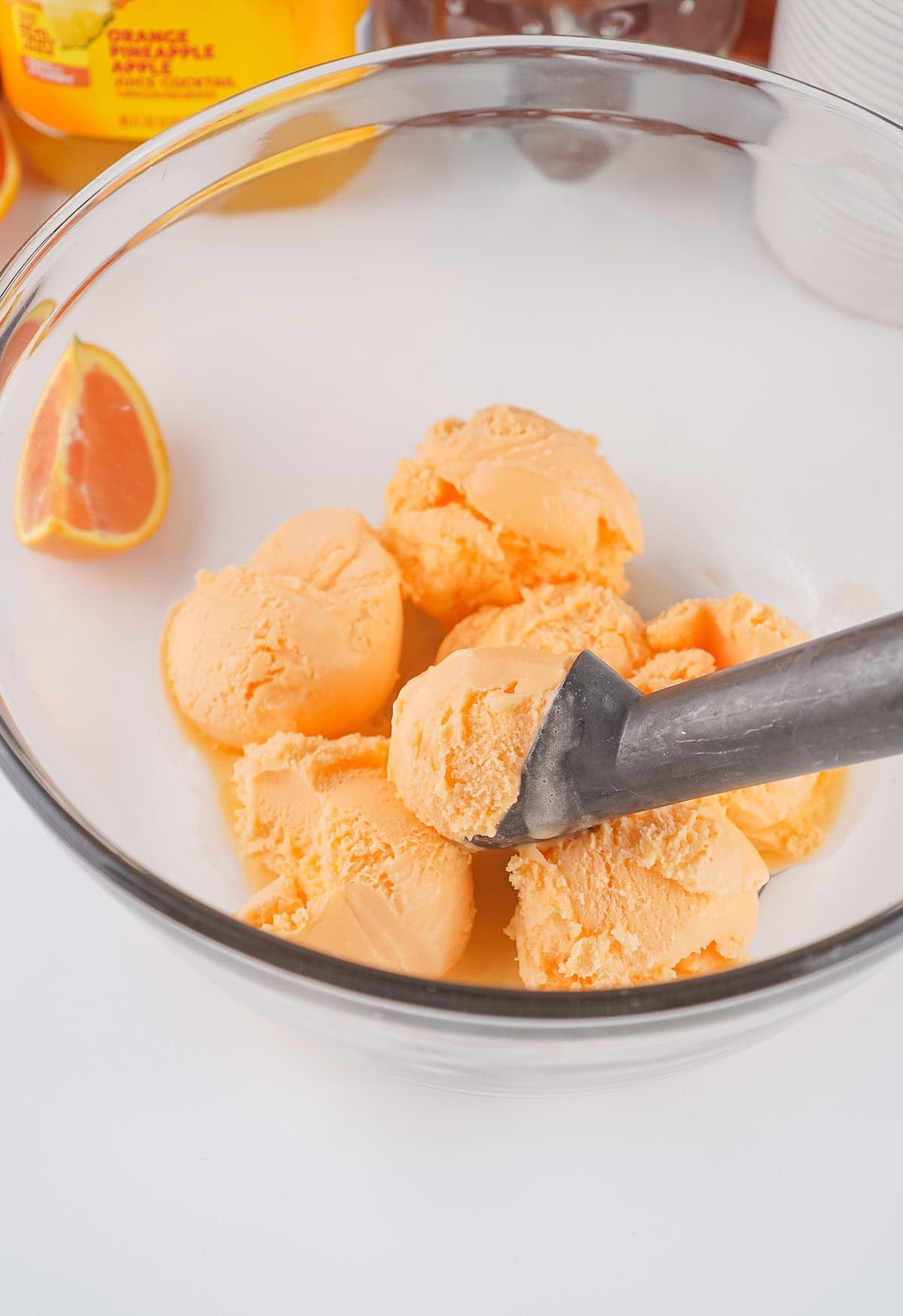 Scoops of orange sherbet in a glass bowl with an ice cream scoop and orange slice nearby.
