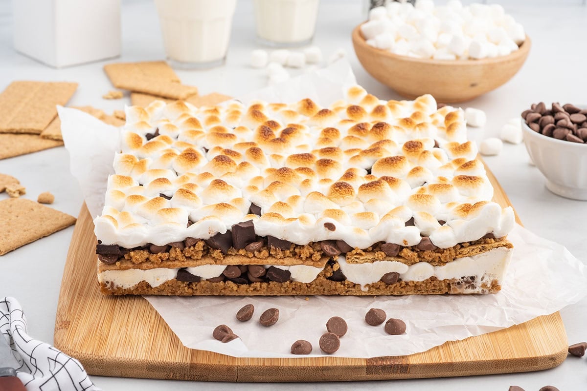 Side view of a large oven-baked s’mores dessert layered with graham crackers, melted chocolate chips, and toasted marshmallows on top, styled with marshmallows and chocolate chips in the background.