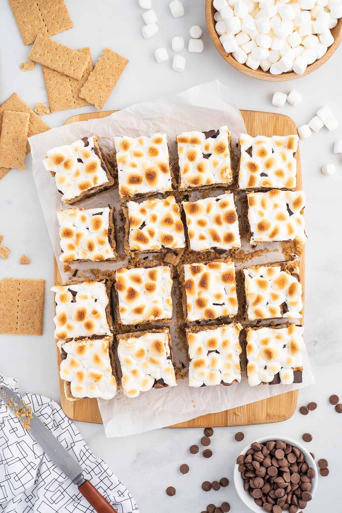 Top-down view of sliced oven-baked s’mores bars with toasted marshmallow topping, graham cracker layers, and melted chocolate, surrounded by marshmallows, graham crackers, and chocolate chips.