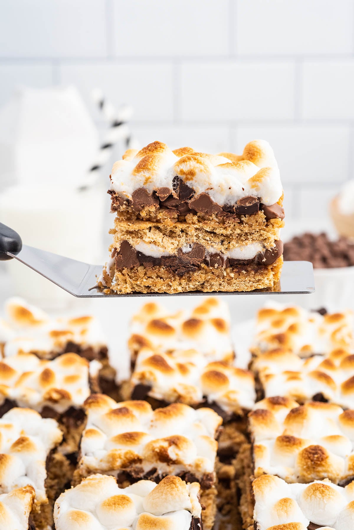 Close-up of a toasted s’mores bar square lifted on a spatula, showing layers of graham crackers, melted chocolate chips, and gooey marshmallows with extra bars blurred in the background.