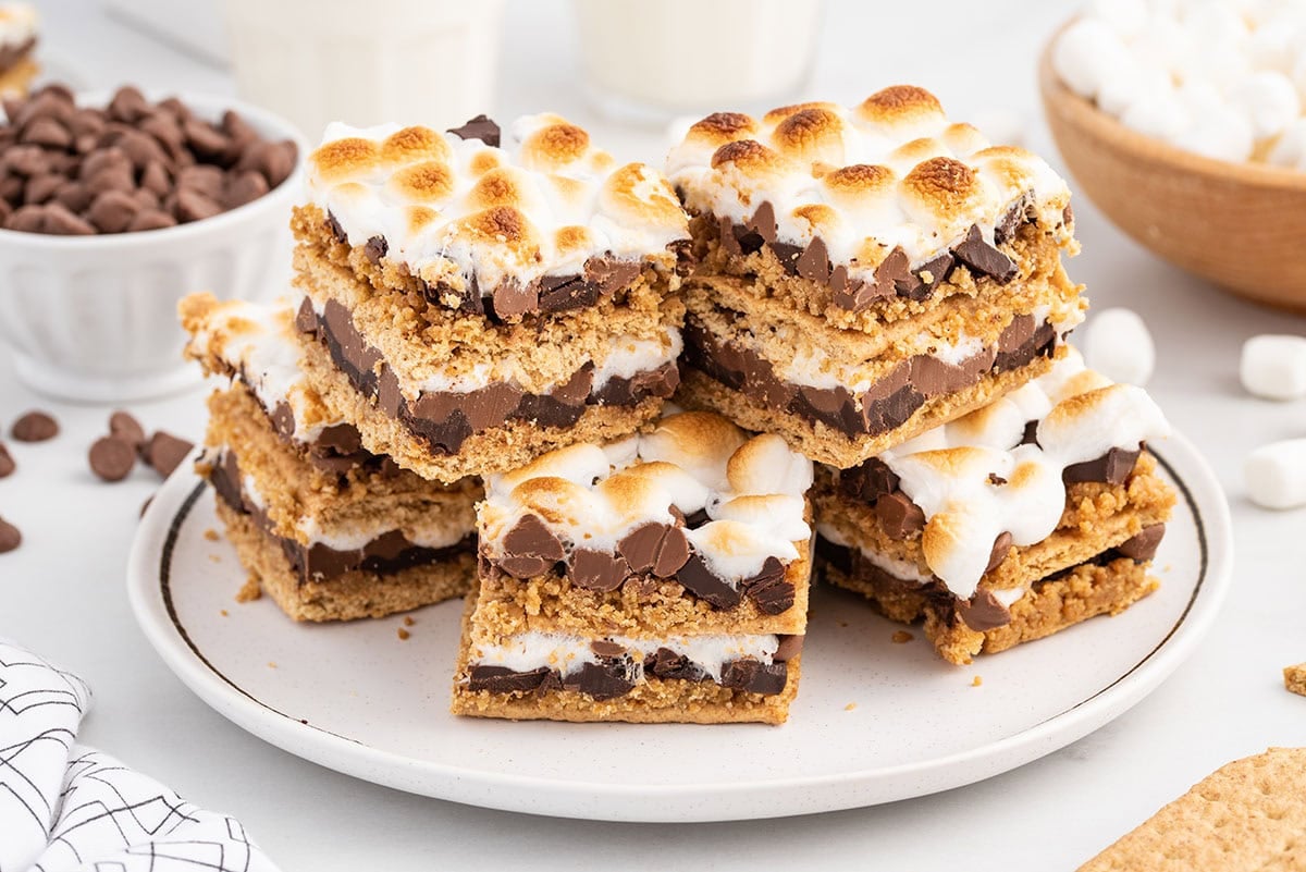 Plate of oven-baked s’mores bars stacked high with layers of graham cracker crust, melted chocolate, and toasted marshmallows, surrounded by chocolate chips and mini marshmallows.