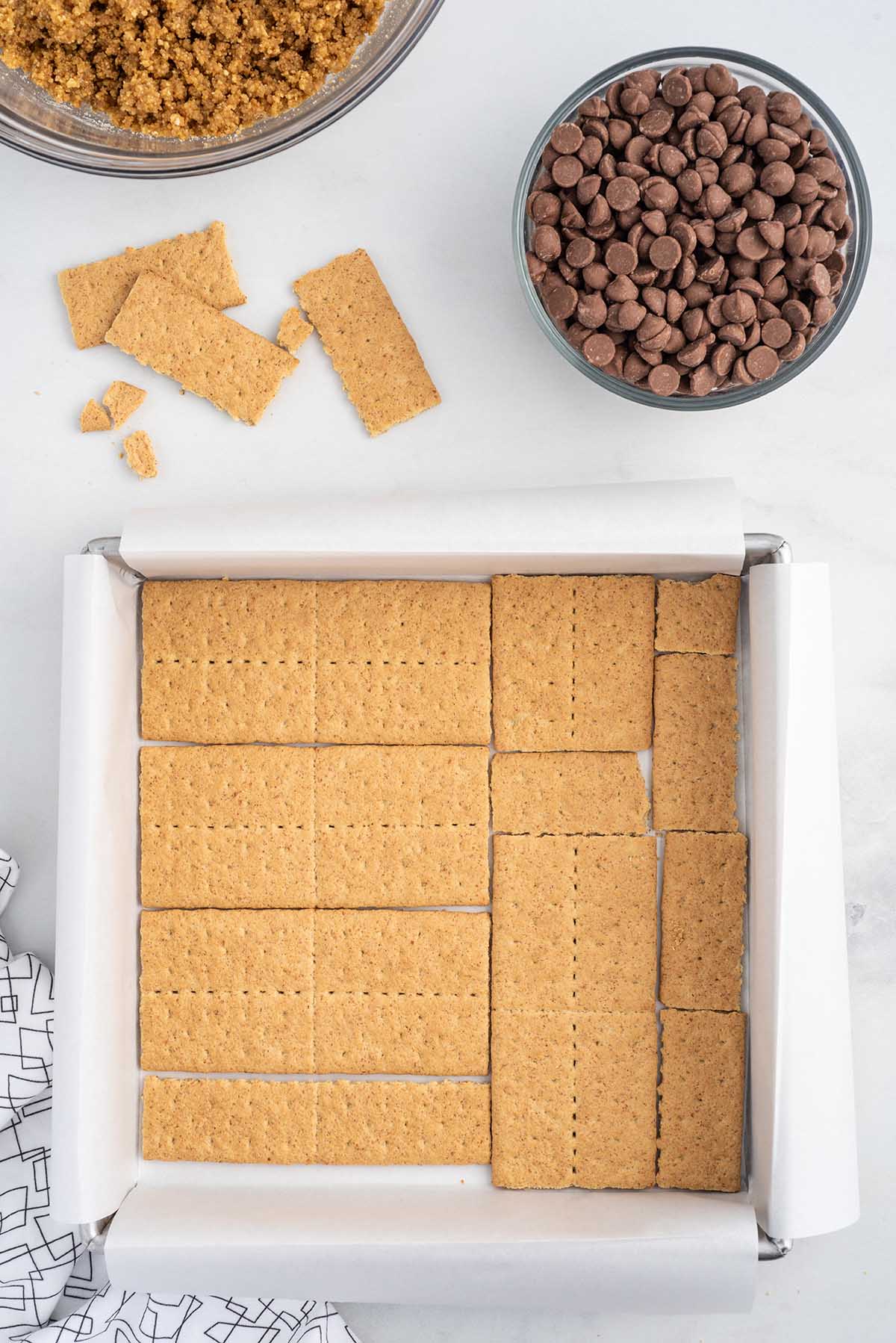 Top-down process shot of graham crackers lined in a parchment-lined baking dish for layered oven-baked s’mores bars, with chocolate chips and graham cracker crumbs nearby.