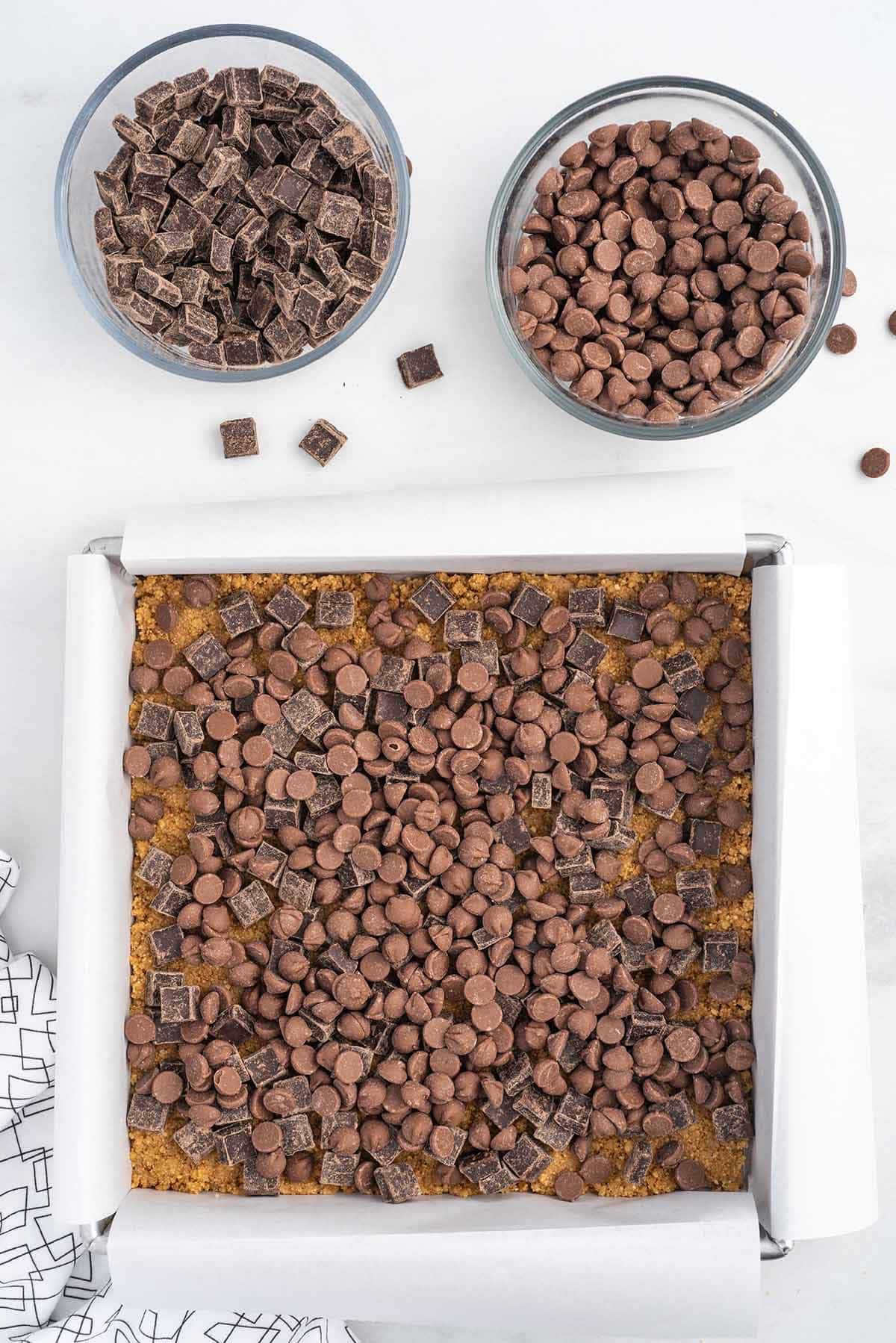 Top-down process photo of a graham cracker crust topped generously with milk chocolate chips and chopped chocolate pieces in a parchment-lined baking pan for oven-baked s’mores bars.