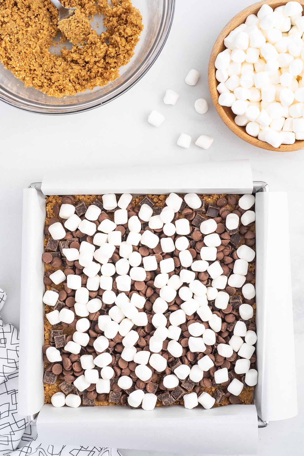 Top-down process shot of mini marshmallows scattered over layers of chocolate chips and chopped chocolate in a parchment-lined baking pan for oven-baked s’mores bars.