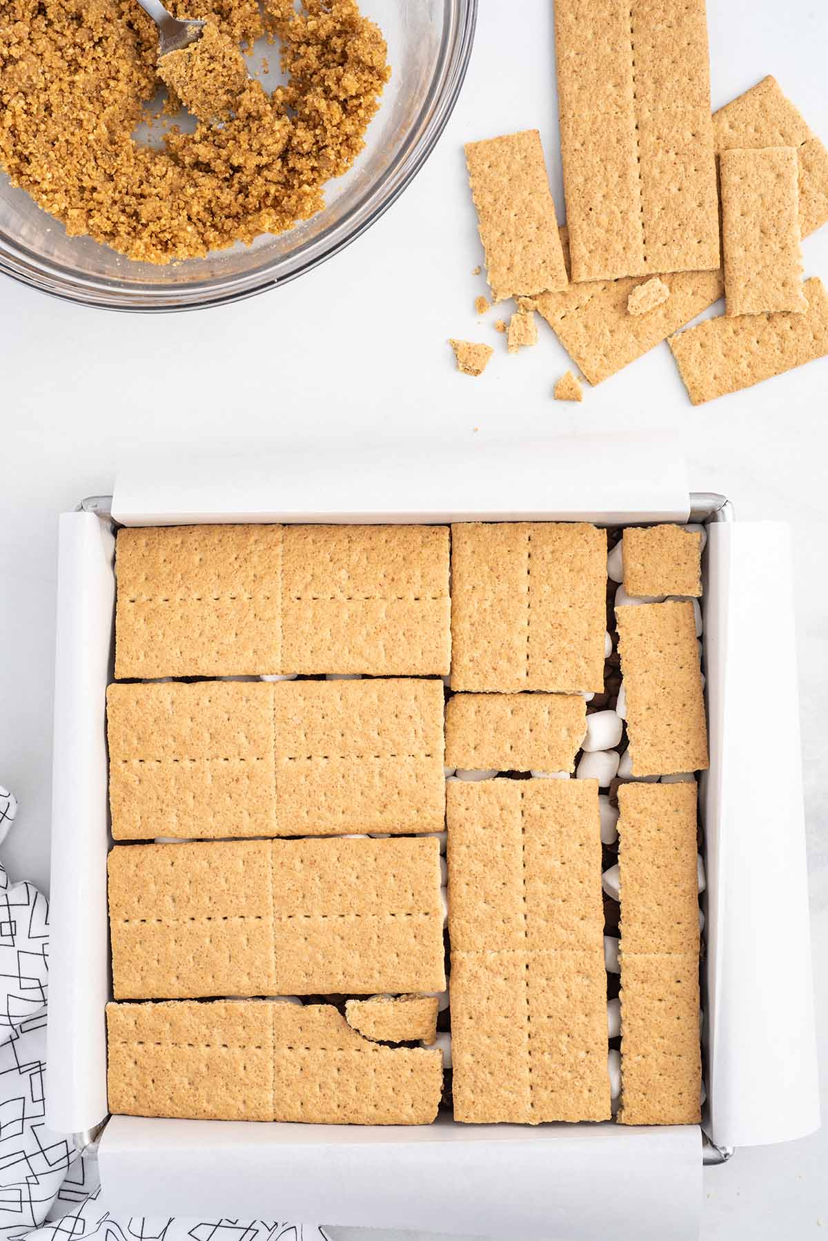 Top-down assembly shot of oven-baked s’mores bars with a final layer of graham crackers arranged over marshmallows and chocolate in a parchment-lined baking pan.