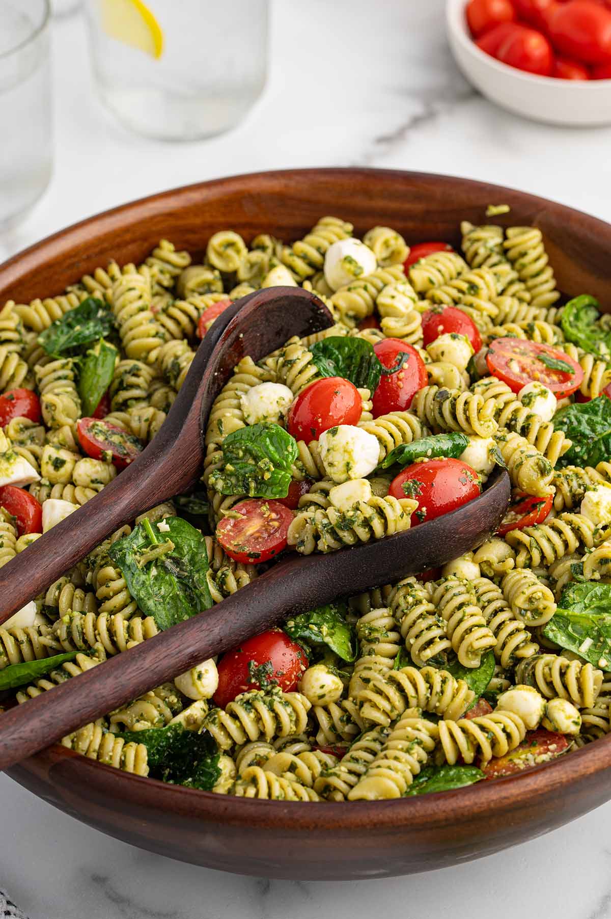 Fresh pesto pasta salad loaded with mozzarella pearls, spinach, and cherry tomatoes in a wooden serving bowl.