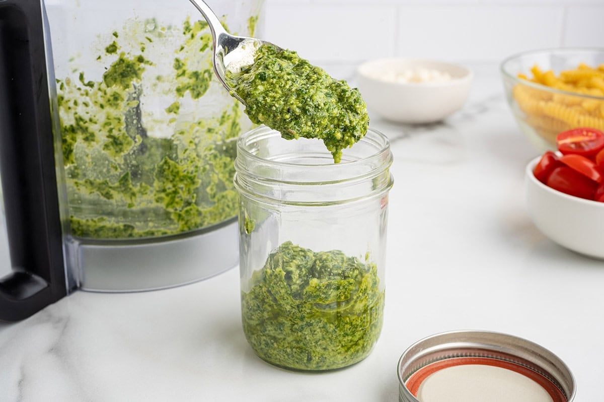 Fresh homemade pesto being spooned into a jar for pesto pasta salad or meal prep.