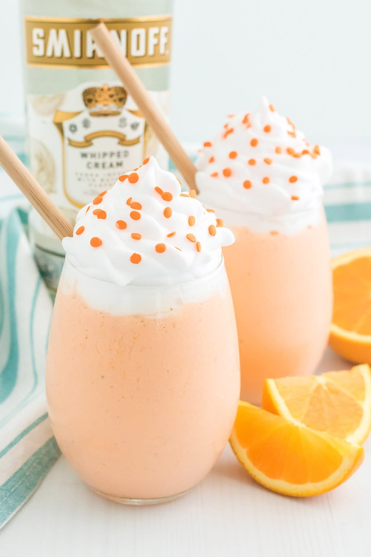 Two Boozy Orange Creamsicle Milkshakes topped with whipped cream and orange sprinkles, with fresh orange slices and a bottle of Smirnoff Whipped Cream vodka nearby.