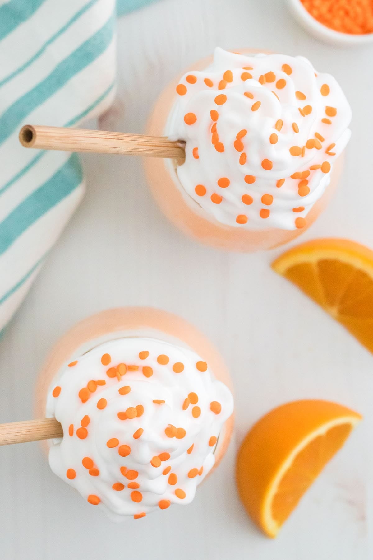 Overhead view of two Orange Creamsicle Milkshakes topped with whipped cream and orange sprinkles, with fresh orange slices alongside.