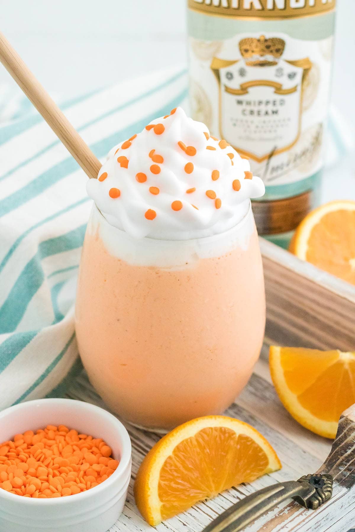 A single Boozy Orange Creamsicle Milkshake topped with whipped cream and orange sprinkles on a wooden tray, with orange slices, a bowl of sprinkles, and Smirnoff Whipped Cream vodka nearby.