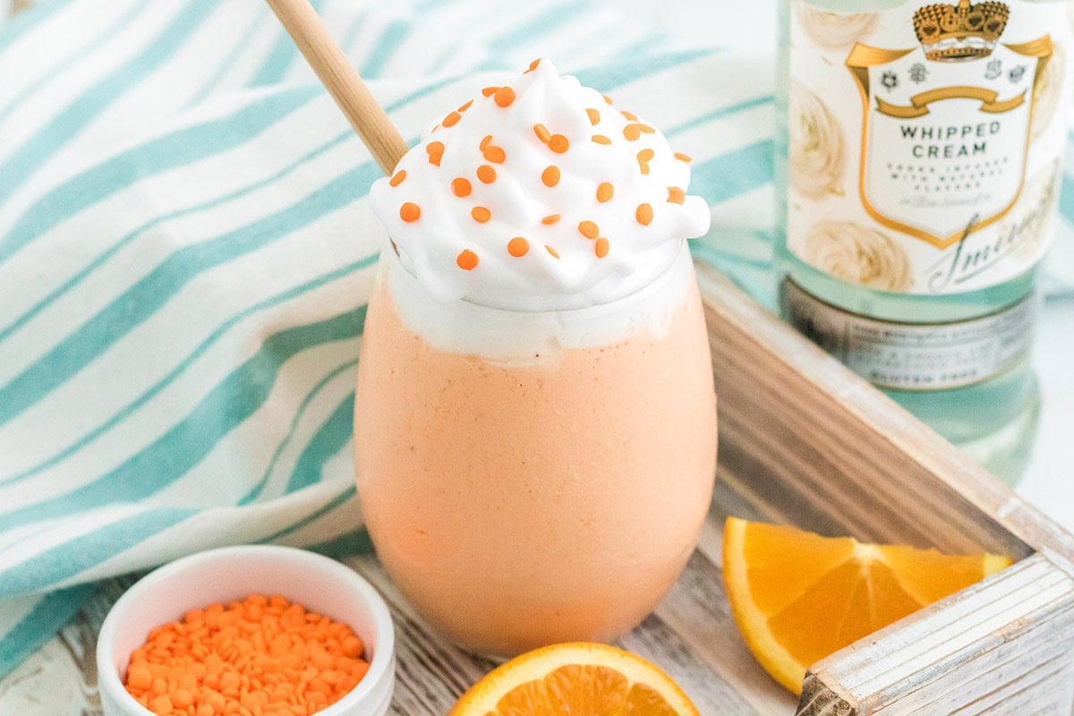 Boozy Orange Creamsicle Milkshake with whipped cream and orange sprinkles on a wooden tray, with orange slices, a bowl of sprinkles, and Smirnoff Whipped Cream vodka in the background.
