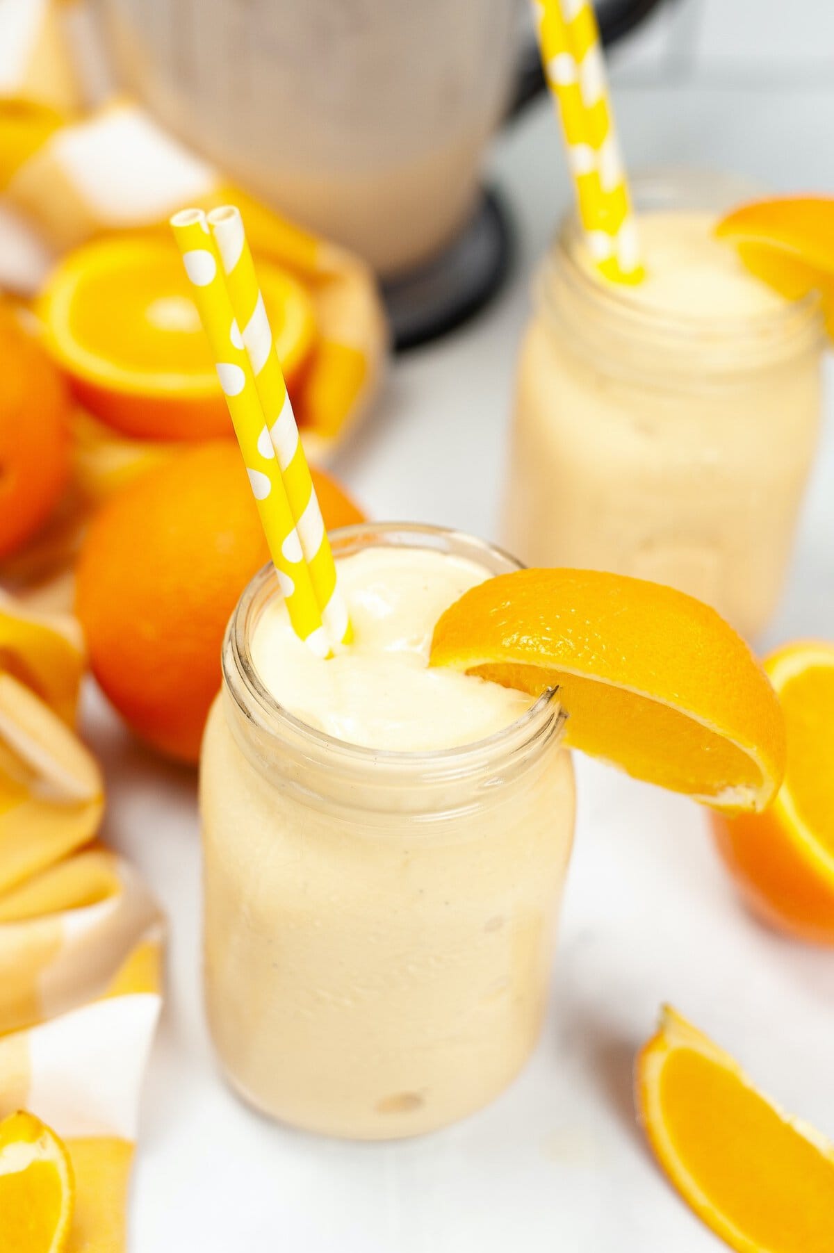 Creamy orange smoothie in a mason jar with an orange slice garnish, surrounded by fresh oranges and yellow-striped straws.