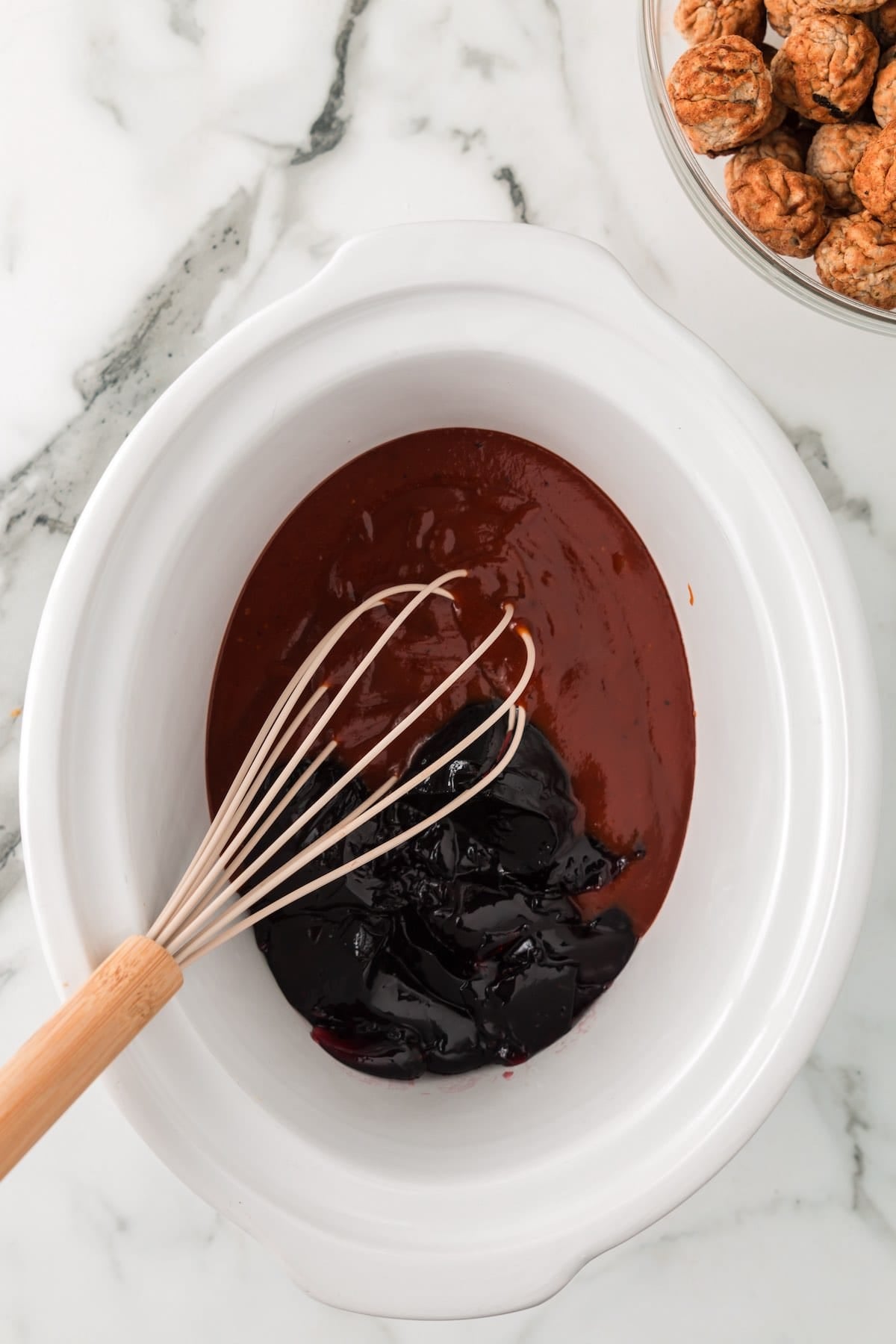 Grape jelly and barbecue sauce being whisked together in a slow cooker for grape jelly meatballs.
