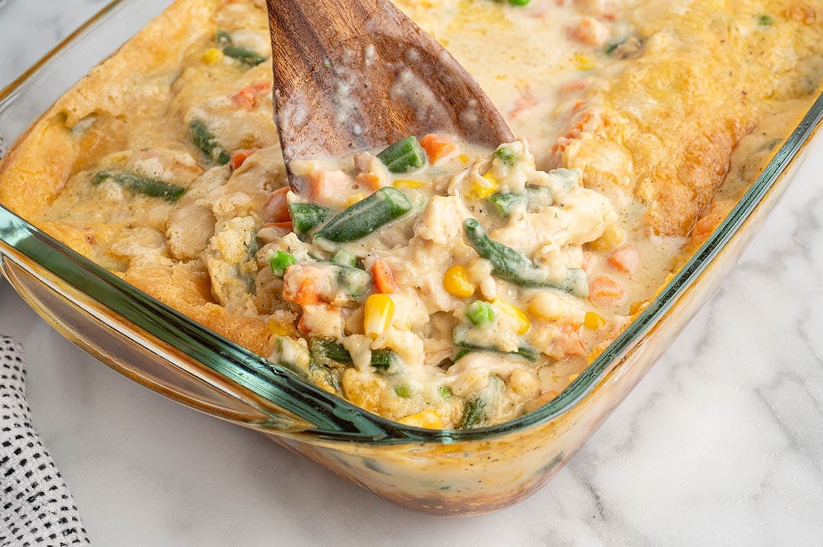 Wooden spoon scooping chicken cobbler with vegetables from a baking dish.