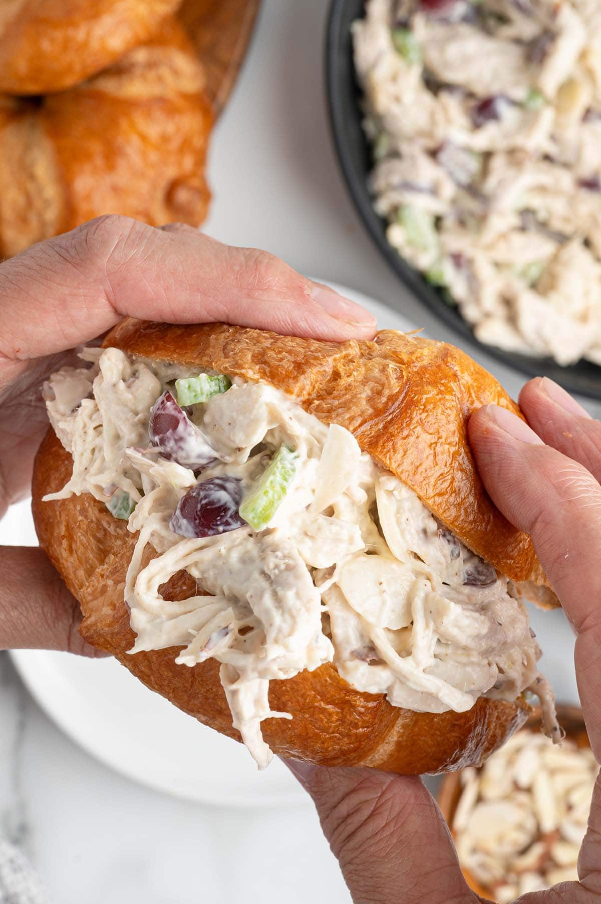 Hands holding a buttery croissant filled with creamy chicken salad, grapes, and celery.