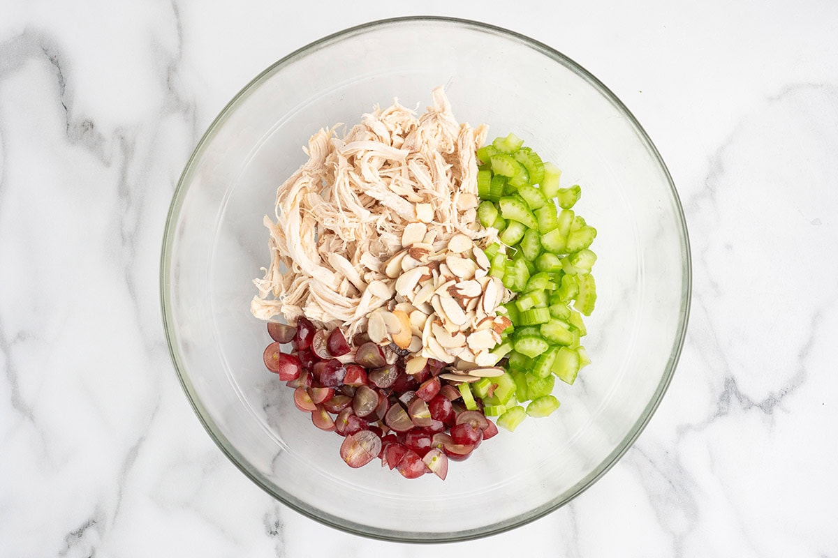 Chicken salad ingredients including shredded chicken, grapes, celery, and sliced almonds in a mixing bowl.