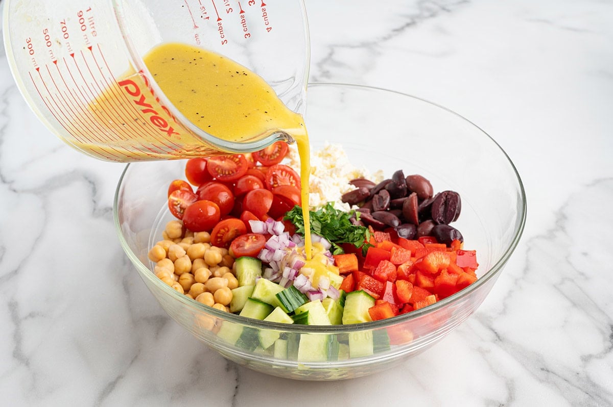 Pouring homemade lemon vinaigrette over chickpeas, cucumber, tomatoes, olives, feta, and vegetables in a large glass mixing bowl.