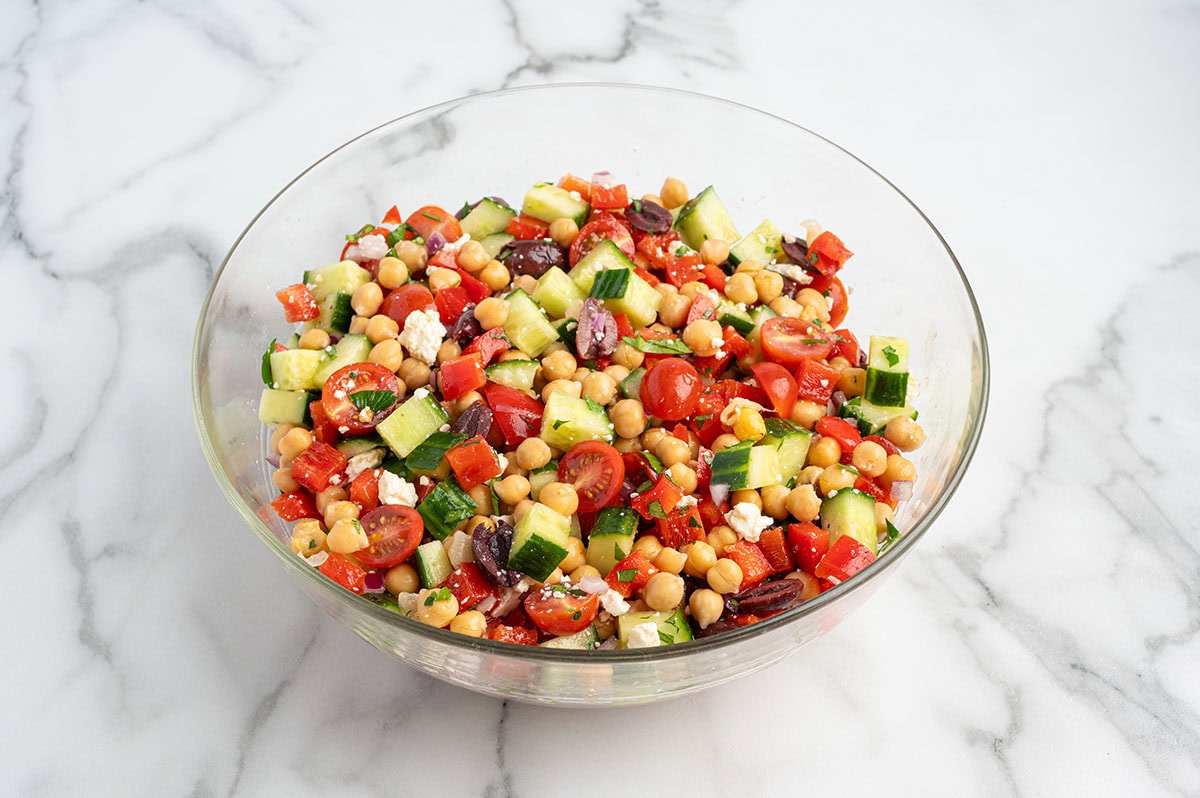 Mediterranean chickpea salad tossed in lemon vinaigrette with cucumbers, tomatoes, red peppers, olives, red onion, parsley, and feta cheese in a glass bowl.