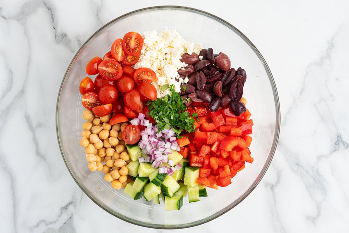 Bowl filled with chickpeas, cucumbers, tomatoes, red bell pepper, olives, feta, red onion, and parsley before mixing.