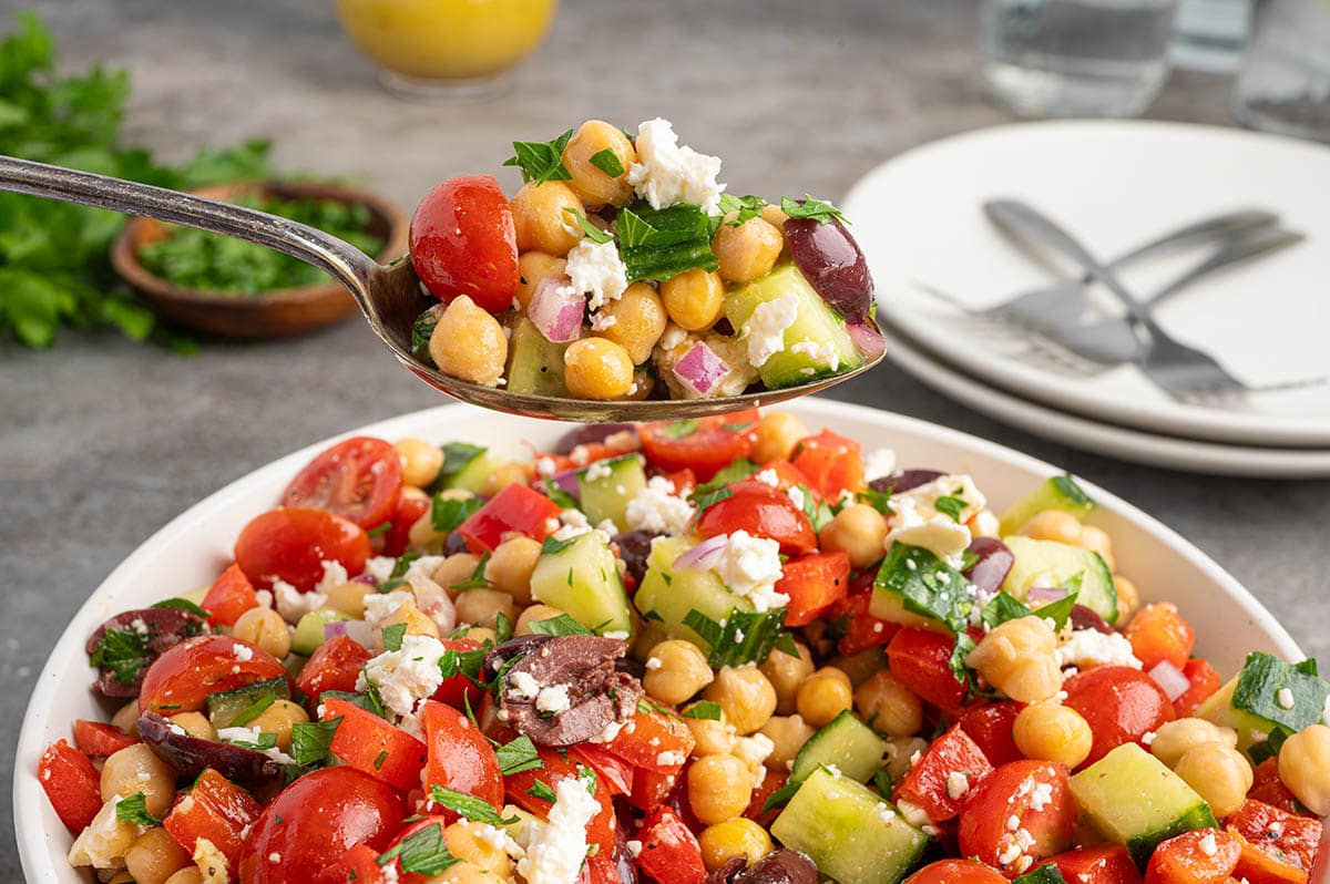 Spoonful of chickpea salad with cucumbers, tomatoes, olives, feta, and herbs lifted above a bowl.