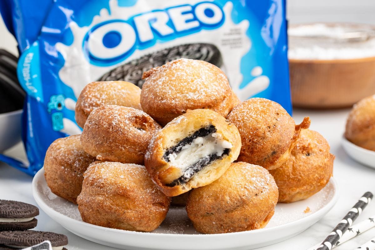 Deep Fried Oreos dusted with powdered sugar piled on a white plate, with one broken open showing the Oreo inside, and a bag of Oreos in the background.