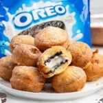 A plate of Deep Fried Oreos dusted with powdered sugar, with one broken open showing the cream filling, and an Oreo bag in the background.
