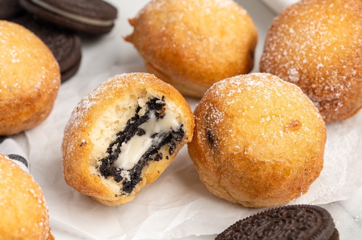 Deep Fried Oreos dusted with powdered sugar in a tray lined with diner paper, with two broken open showing the cream filling, alongside glasses of milk and Oreo cookies.