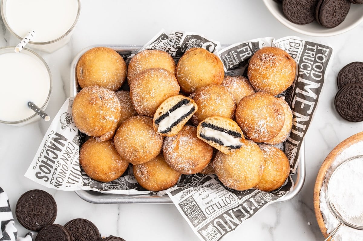 Deep Fried Oreos dusted with powdered sugar in a tray lined with diner paper, with two broken open showing the cream filling, alongside glasses of milk and Oreo cookies.