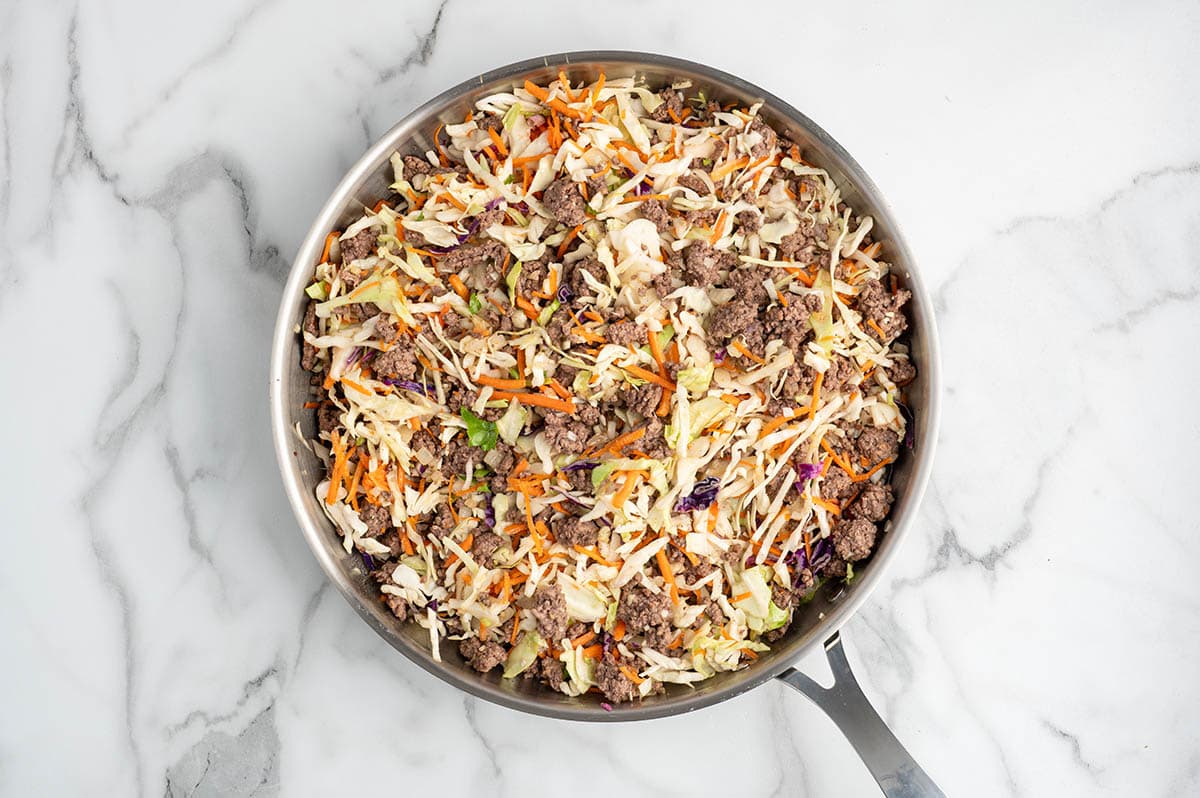 Coleslaw mix and browned ground beef cooking together in a skillet for egg roll in a bowl.