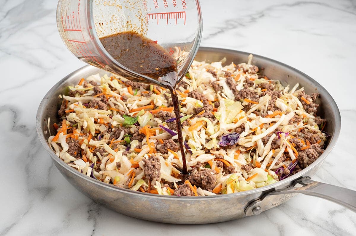 Sauce being poured into a skillet with ground beef and coleslaw mix for egg roll in a bowl.