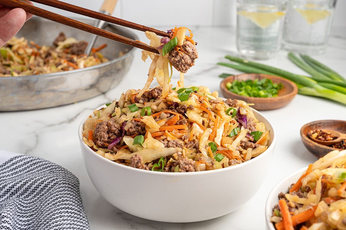 Chopsticks lifting a bite of egg roll in a bowl with ground beef, cabbage, carrots, and green onions from a white bowl.