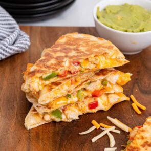 Stacked chicken fajita quesadilla wedges with melted cheese and peppers on a wooden board next to a bowl of guacamole.
