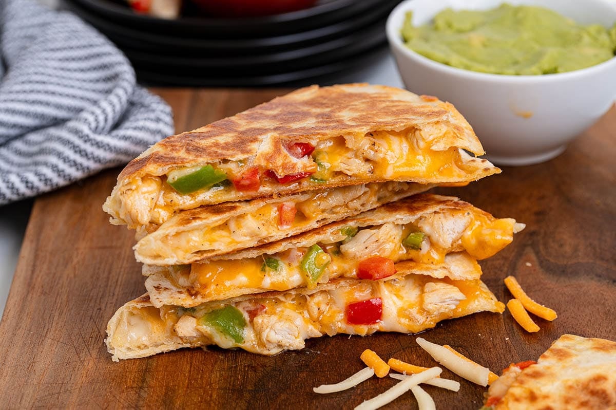 Stacked chicken fajita quesadilla wedges with melted cheese, peppers, and chicken on a wooden board with guacamole.