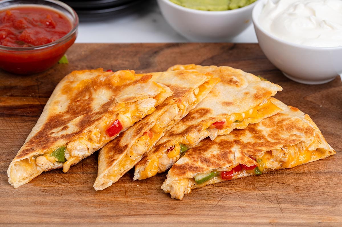 Chicken fajita quesadilla wedges with melted cheese and peppers on a wooden board, served with salsa, guacamole, and sour cream.