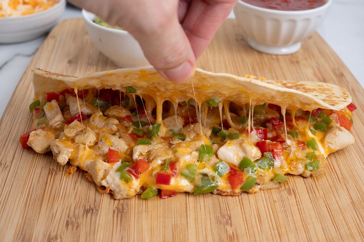 Hand lifting the top of a chicken fajita quesadilla to reveal melted cheese, chicken, and peppers.