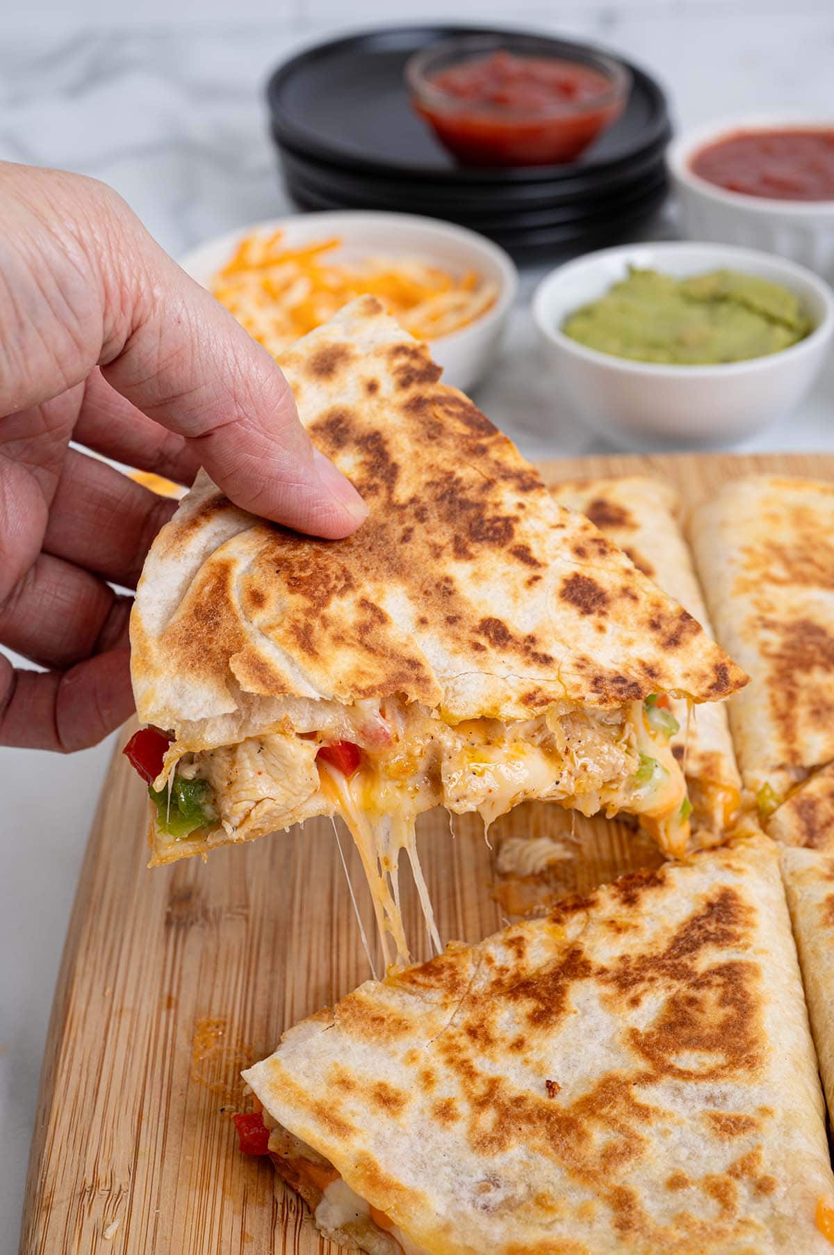Hand lifting a cheesy chicken fajita quesadilla wedge with melted cheese stretching, served with salsa, guacamole, and shredded cheese.