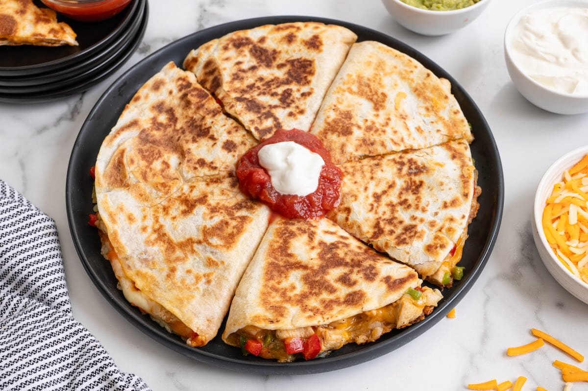 Chicken fajita quesadilla cut into wedges on a black plate, topped with salsa and sour cream, served with shredded cheese, guacamole, and sour cream.