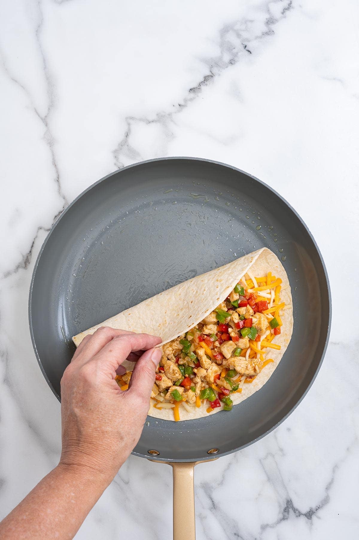 Hand folding a tortilla over cheese and chicken fajita filling in a skillet.