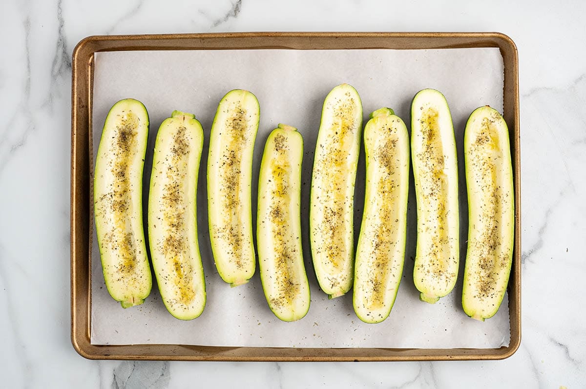 Hollowed zucchini halves arranged on a parchment-lined baking sheet, brushed with oil and seasoned with herbs and black pepper before baking.