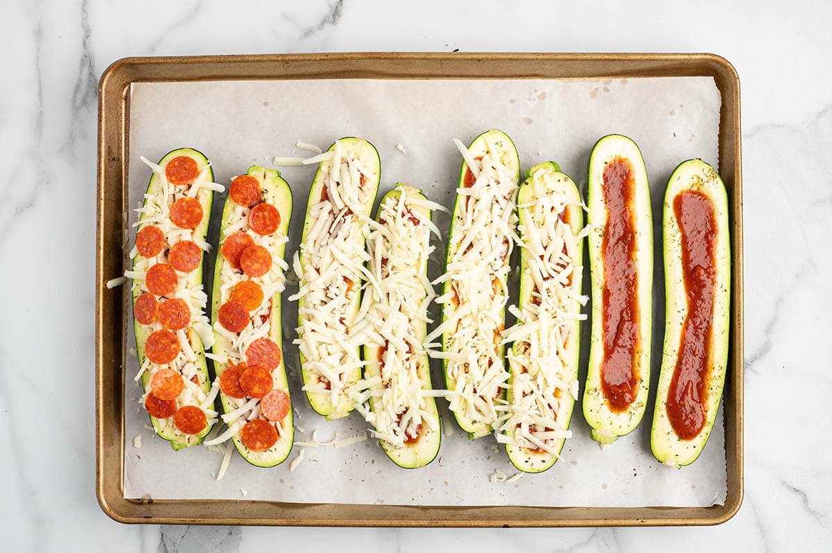 Zucchini pizza boats lined up on a baking sheet, showing different stages of assembly with pizza sauce, shredded mozzarella, and mini pepperoni slices.