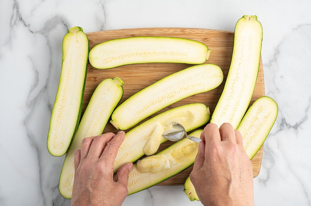 Hands scooping the centers from halved zucchini to create zucchini boats on a cutting board.