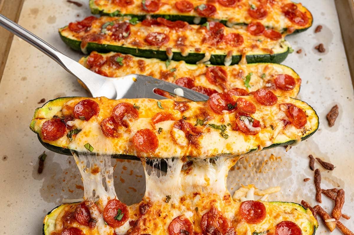 A spatula lifting a pizza zucchini boat with melted cheese stretching from the pan, topped with mini pepperoni and basil.