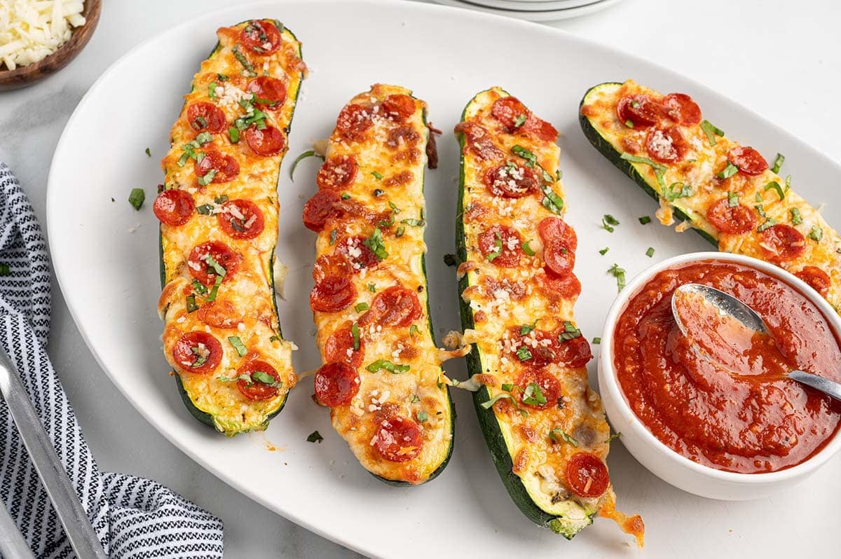 Pizza zucchini boats with melted cheese, mini pepperoni, and basil on a white platter with a side of marinara sauce.