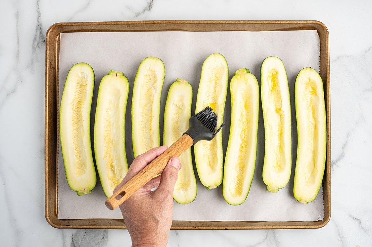 Hand brushing oil onto hollowed zucchini halves arranged on a parchment-lined baking sheet, preparing them for zucchini pizza boats.