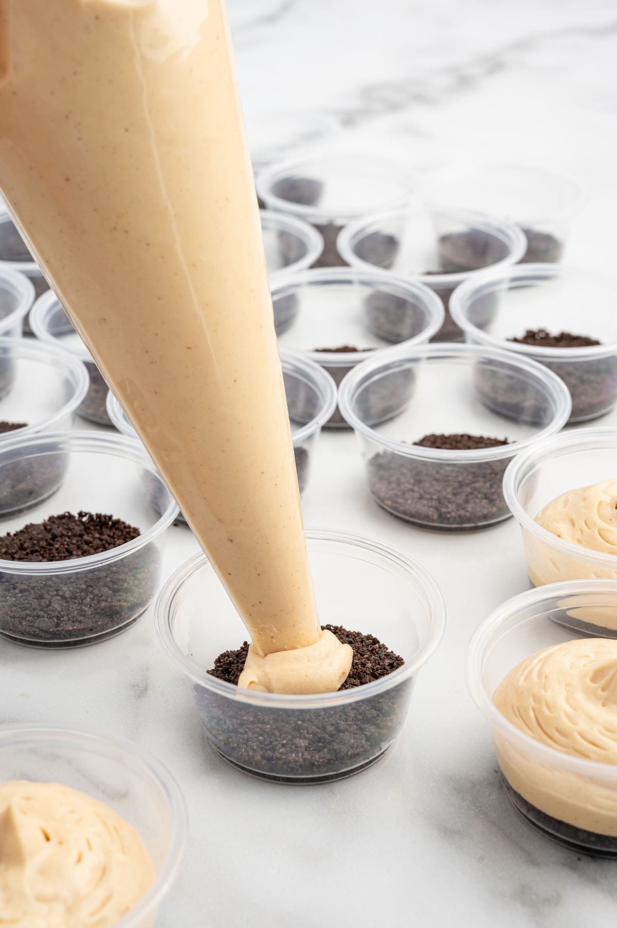 Peanut butter cheesecake filling being piped into dessert cups over a chocolate cookie crumb crust.