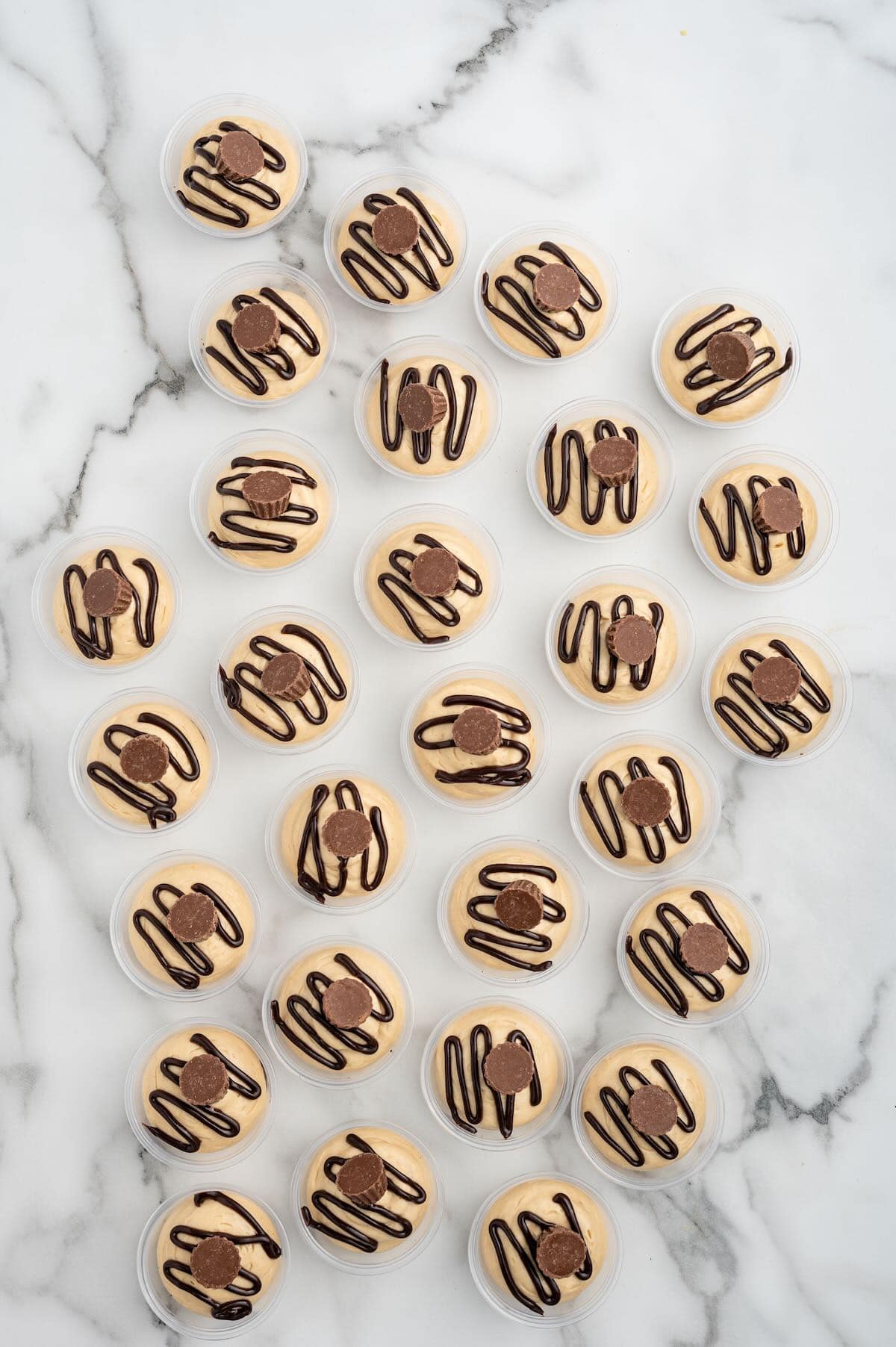 Individual peanut butter cheesecake dessert cups topped with a chocolate drizzle and peanut butter cups, arranged on a marble surface.